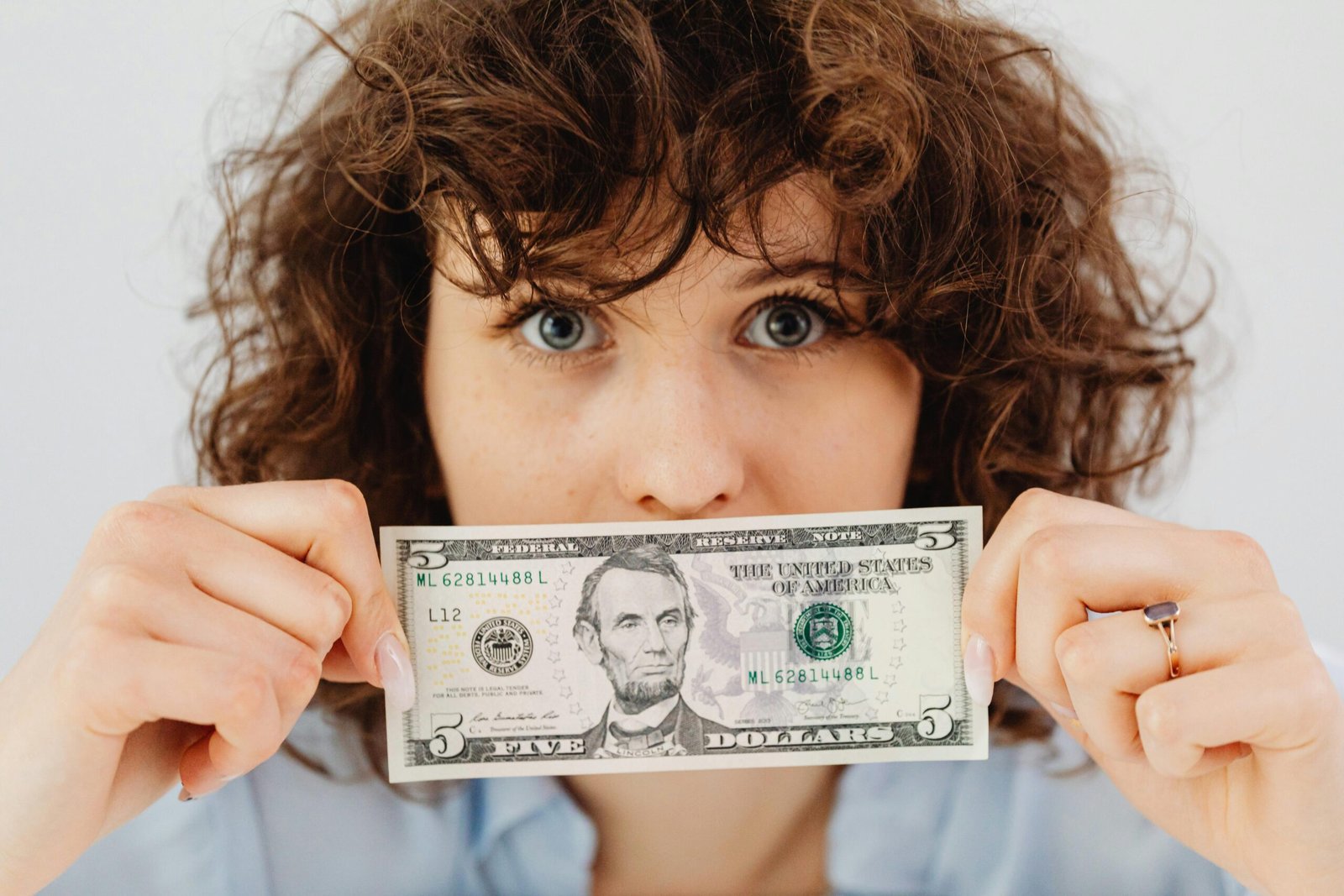 Close-up of a woman with curly hair holding a five dollar bill in front of her face.